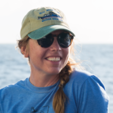 Amy Gartman wearing a light blue shirt looks off camera to the right smiling wearing large sunglasses and a tan and sage green baseball hat