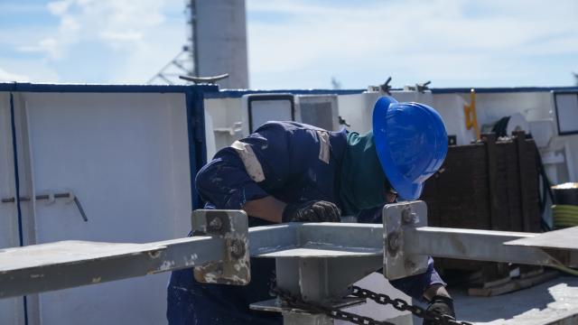 A man in a navy jumpsuit with grey shoulder strips and a light blue hard hat kneeling down besides a metal structure. The man is building the grey structure on the ship's stern deck. 