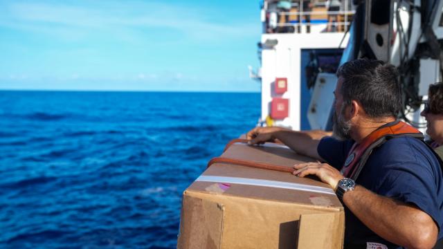 Image shows a man with short black hair weary a navy blue shirt and an orange life preserver. The man is holding a cardboard box over the side of a ship. The cardboard box had two red ropes tied around it. Behind the man with the box, is the white paint of the ship. In the background there is light blue sky and dark blue ocean.  