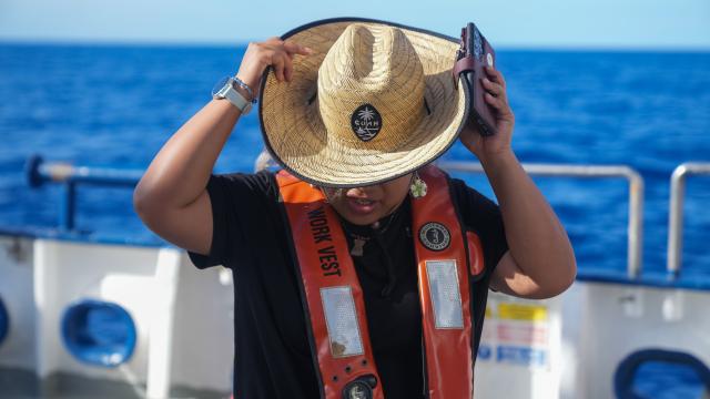A young woman wearing a black shirt and orange life preserver placing a large straw hat on her head. The center of the hat has the Guam seal. Behind her is the outline of the ships stern deck and the dark blue ocean and light blue sky. 