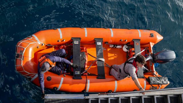A top down image of two men on an orange life boat that has reflective grey stripes along each of the sides. The man on the right is standing in the boat wearing a grey jumpsuit and glasses. The man on the left is sitting in the boat wearing a navy jumpsuit and navy hat. Both men are wearing orange life preservers. The boat is alongside a grey staircase.  
