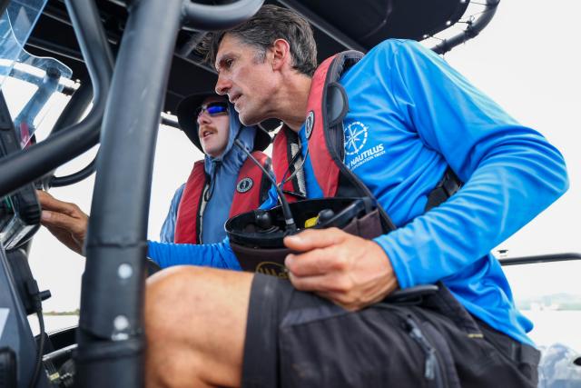 Picture of two men wearing blue shirts with red life preservers sitting inside of a small boat. 