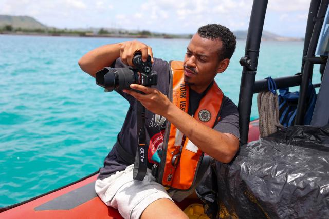 A man wearing a black shirt and white shorts holding a camera and taking a picture. The man is sitting in a small red boat wearing an orange life preserver. 
