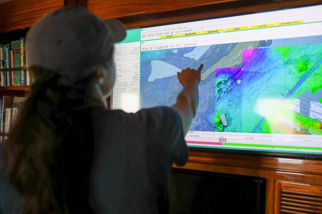A picture of a woman with brown wavy hair wearing a grey shirt and grey hat. The picture shows the back of her head while she points at at TV screen. The TV screen features a colorful bathymetric map of the seafloor.  