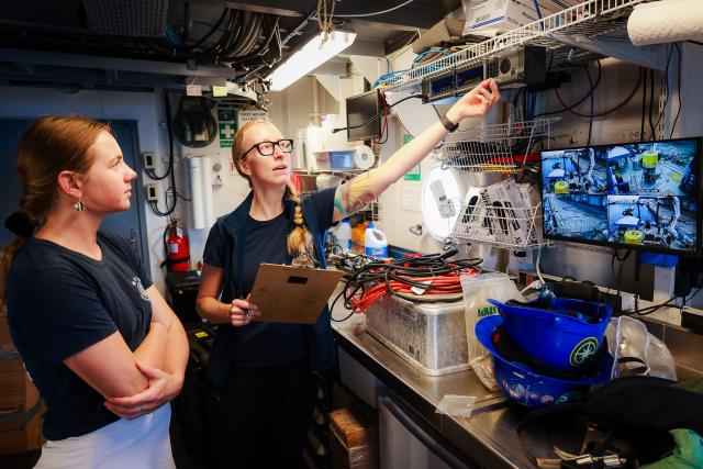 Two women stand in a workspace aboard the E/V Nautilus. The woman on the right, holding a clipboard, points toward a shelf with electronic equipment and cables. She has glasses, a long braid, and a visible arm tattoo. The woman on the left listens attentively with arms crossed. The space is filled with coiled wires, a monitor displaying multiple camera views from the ship’s deck, and two blue hard hats resting on a metal counter. 