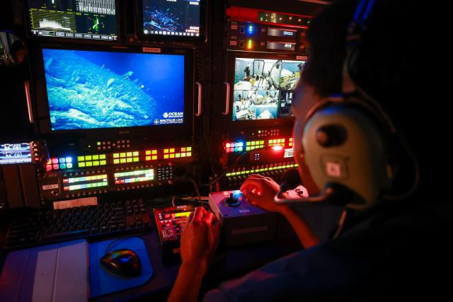 A person wearing a headset operates a control station aboard the E/V Nautilus. The dimly lit room glows with multicolored buttons and display screens. The central monitor shows a live feed of the ocean floor captured by an ROV (Remotely Operated Vehicle), revealing rocky terrain and marine life. Additional monitors display vessel footage and system data. The scene captures the high-tech coordination behind deep-sea exploration, emphasizing the precision and teamwork required to bring real-time ocean science