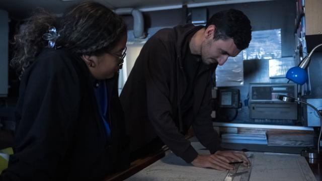 Two people stand closely together in the ships bridge , looking down at a large nautical chart spread across the table. The person on the left, with curly hair tied back and wearing glasses, watches attentively. The person on the right, with short dark hair leans over the map while using a ruler and pencil to plot a course. The room is filled with navigational equipment, documents, and a small blue desk lamp. 