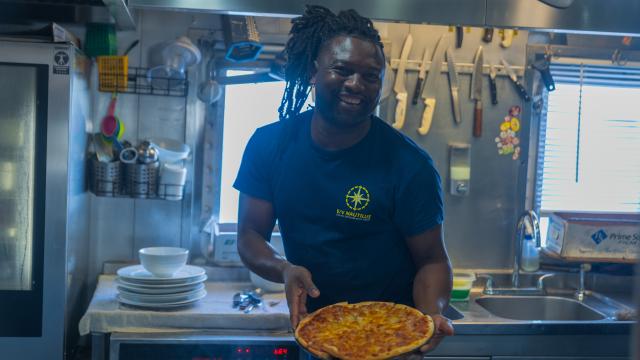 A smiling person wearing a navy blue E/V Nautilus T-shirt stands in a stainless steel ship galley, holding a freshly baked cheese pizza with both hands. Behind them, kitchen utensils, including a variety of knives, are neatly mounted on a wall rack. Stacked plates, bowls, and cooking tools are organized around the well-lit space. Sunlight shines through a window, casting a cheerful glow on the scene.