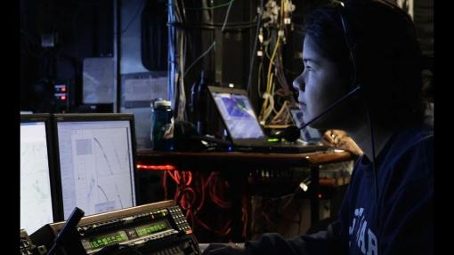 Asian woman sits in dark control van looking at computer screens