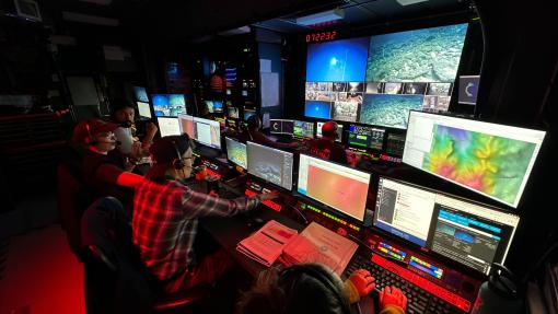 Image of a darkened control room with the team looking at large computer monitors showing the seafloor